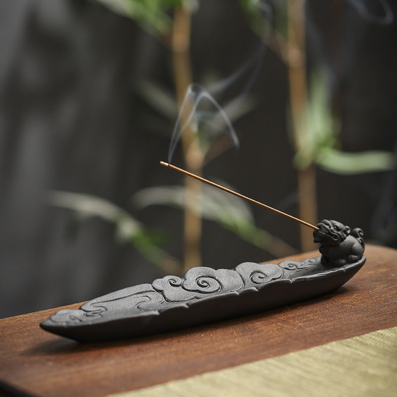 Black ceramic incense stick holder on a dark wood surface with soft-focus greenery